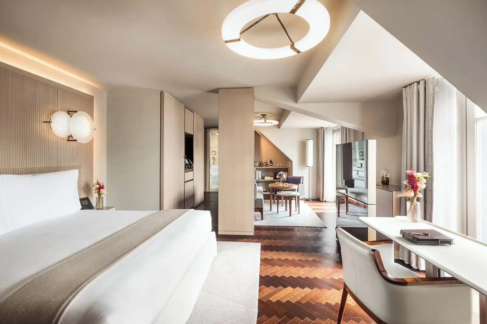 Parisian Suite at Mandarin Oriental Lutetia, Paris: a double bed against a pale fluted headboard and a writing desk under a sloping ceiling, open to a sitting and dining area beyond