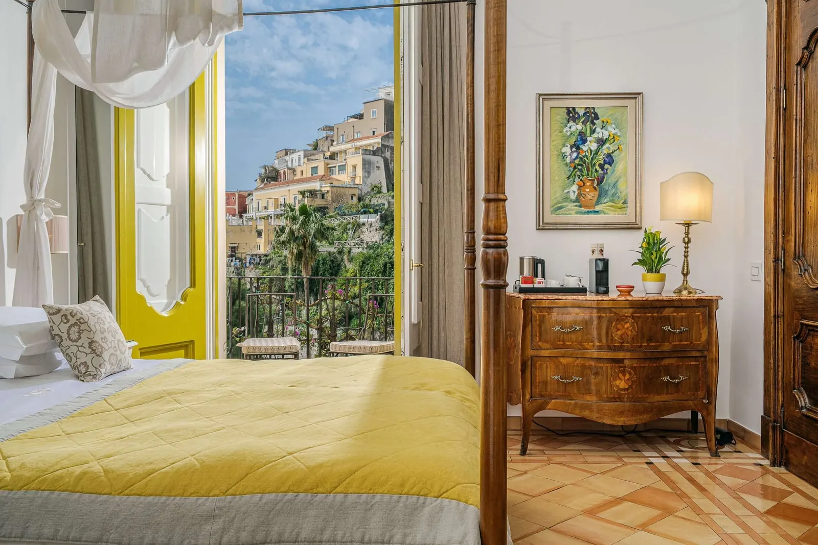 Deluxe Sea View im Palazzo Murat: das Fußende eines Himmelbetts neben einer antiken Kommode, mit gelben Fensterläden, die sich zu einem Balkon öffnen