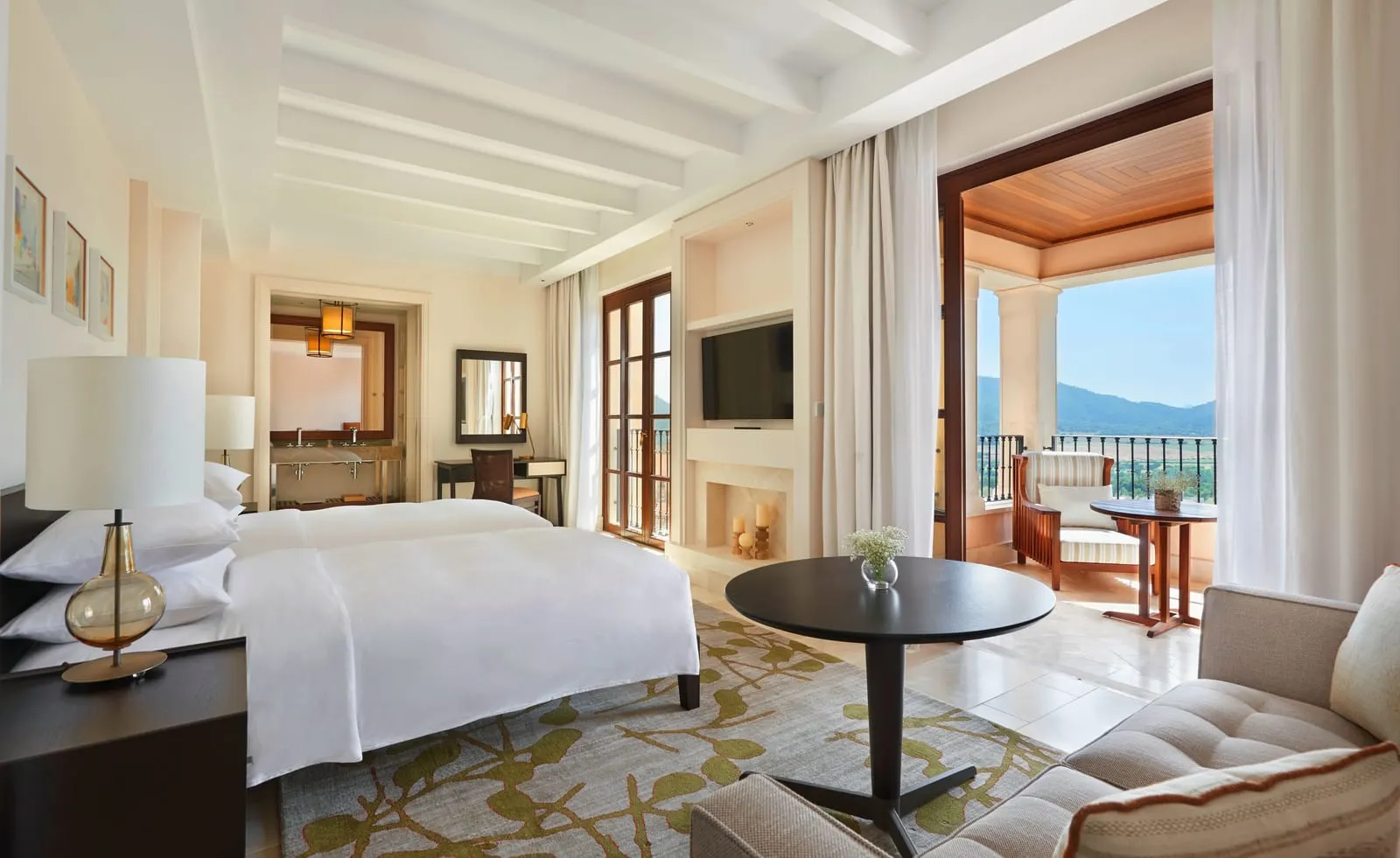 Junior Suite Valley View at Cap Vermell Grand Hotel: a white bed under pale beams, a sofa and round table, and an open door to a terrace with hills beyond