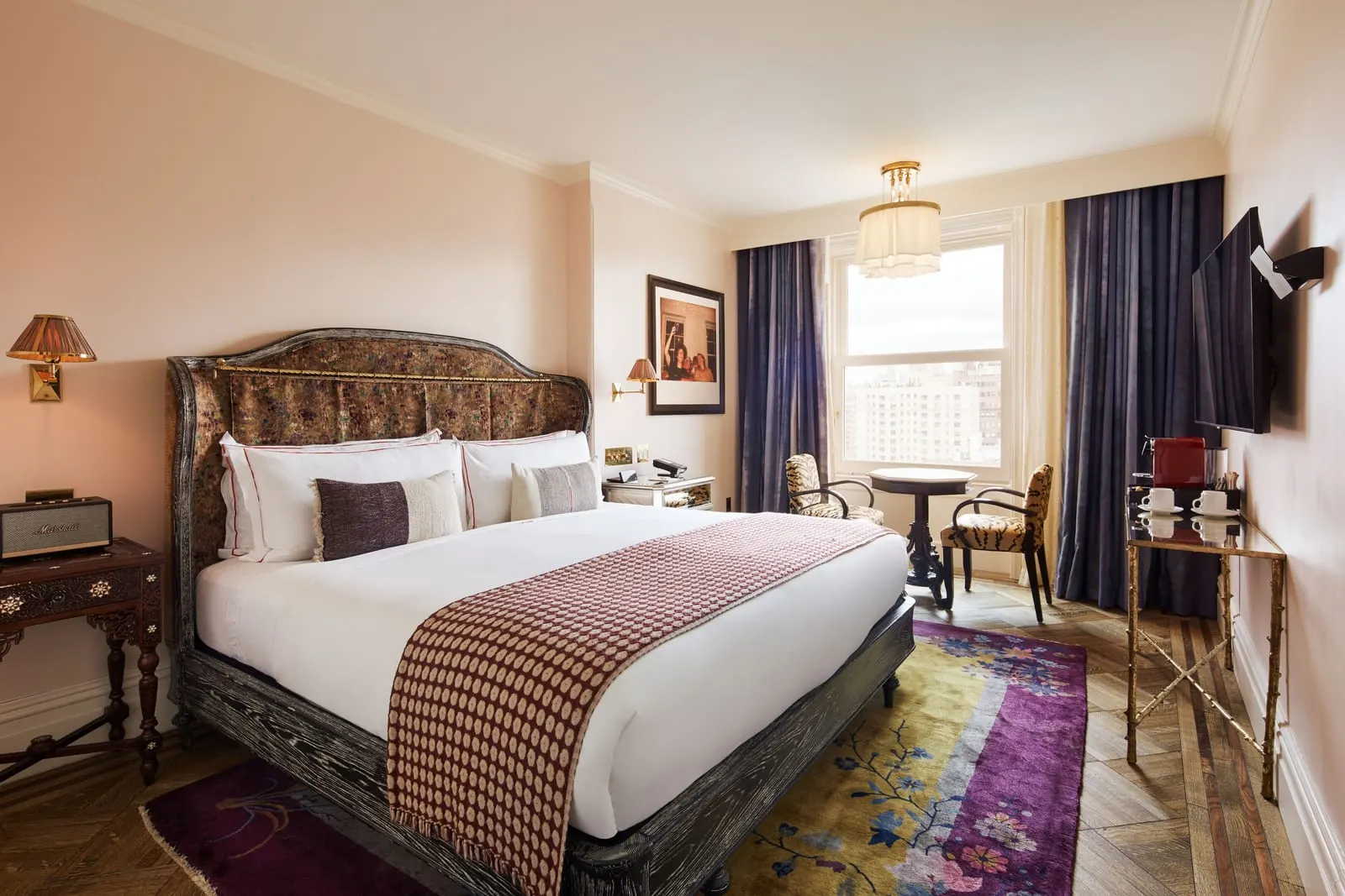 The Chelsea at Hotel Chelsea: a bed with a patterned velvet headboard, a small round table with two tiger-print chairs by a tall window, and a coffee station on a gold side table