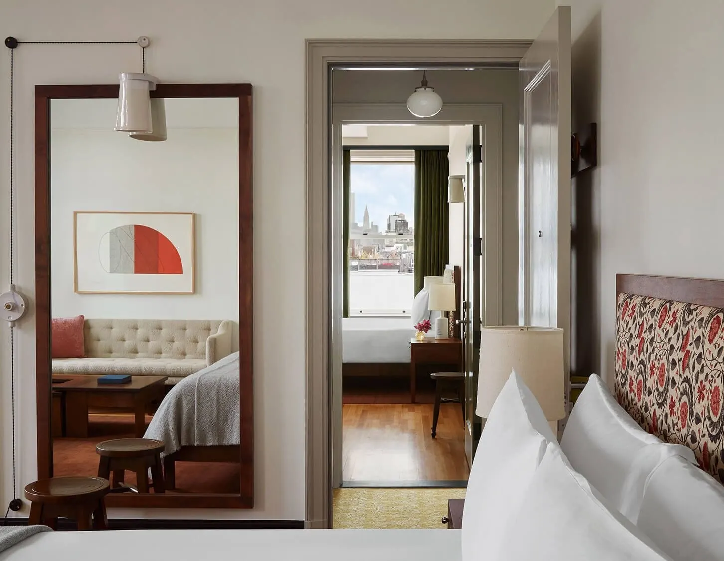 Two Bedroom Supreme Suite at Nine Orchard: a bed with a patterned headboard beside a tall mirror reflecting a sofa, with a doorway through to a second room with another bed