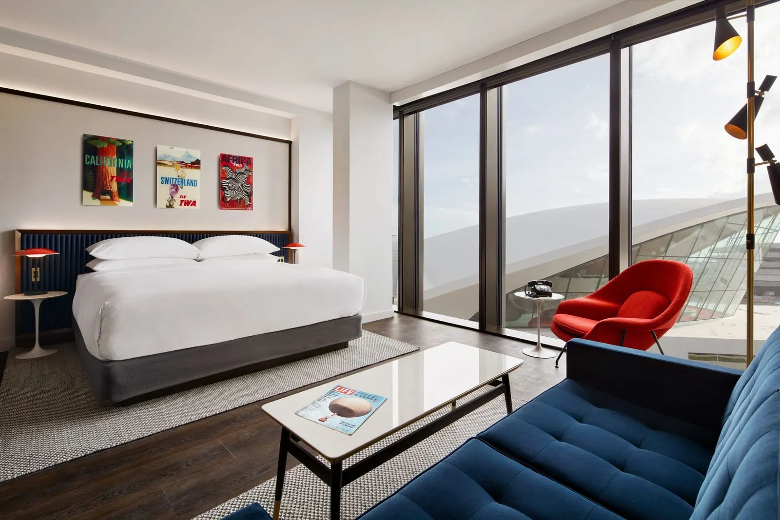 Executive King Suite With Historic TWA View at TWA Hotel: a king bed and a blue sofa facing floor-to-ceiling windows, with the curved white roof of the TWA Flight Center outside and a red armchair beside a rotary phone