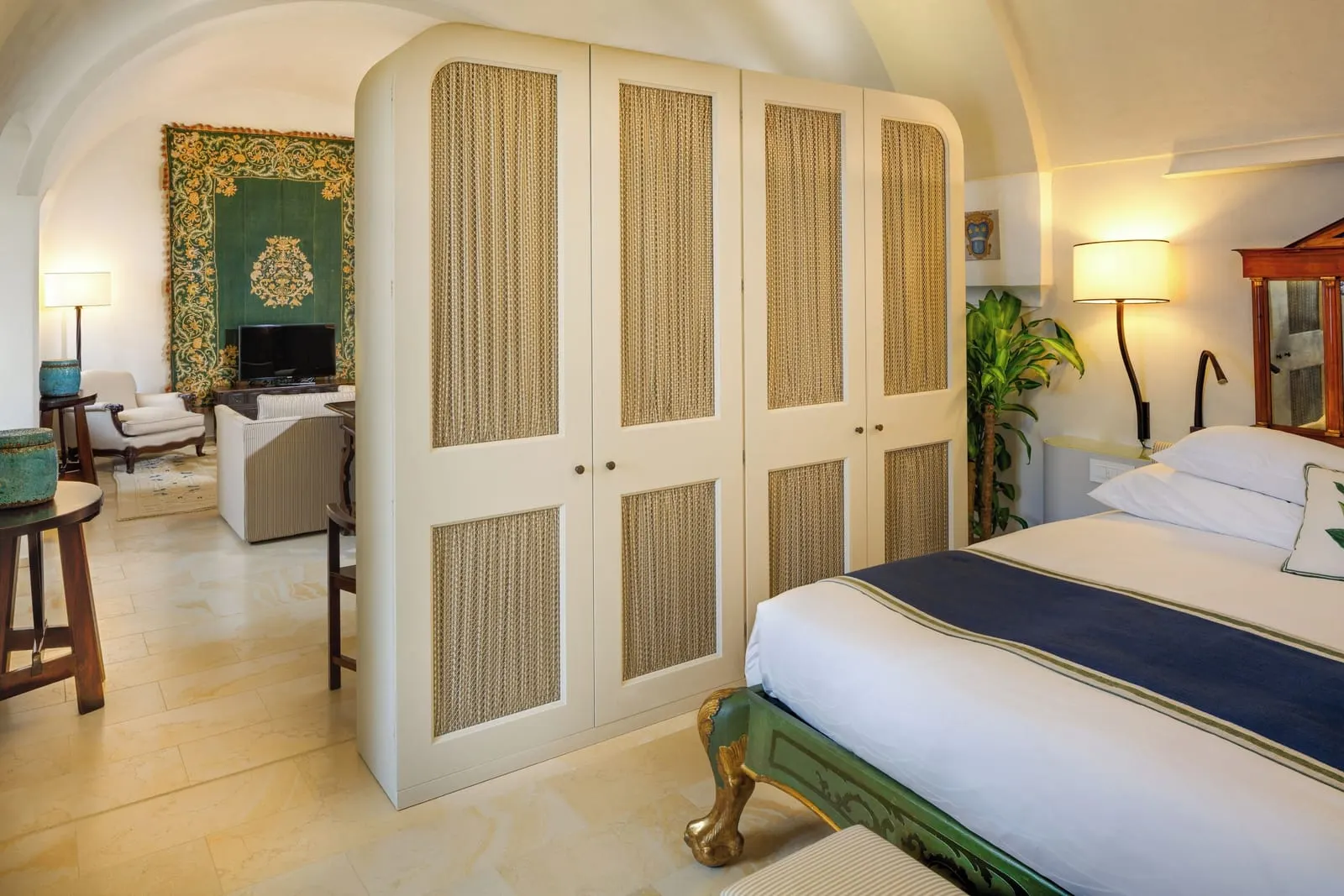 Deluxe Open Space Suite at Monastero Santa Rosa: a king bed beside a tall white wardrobe, with the sitting area and a green tapestry beyond in one open room.