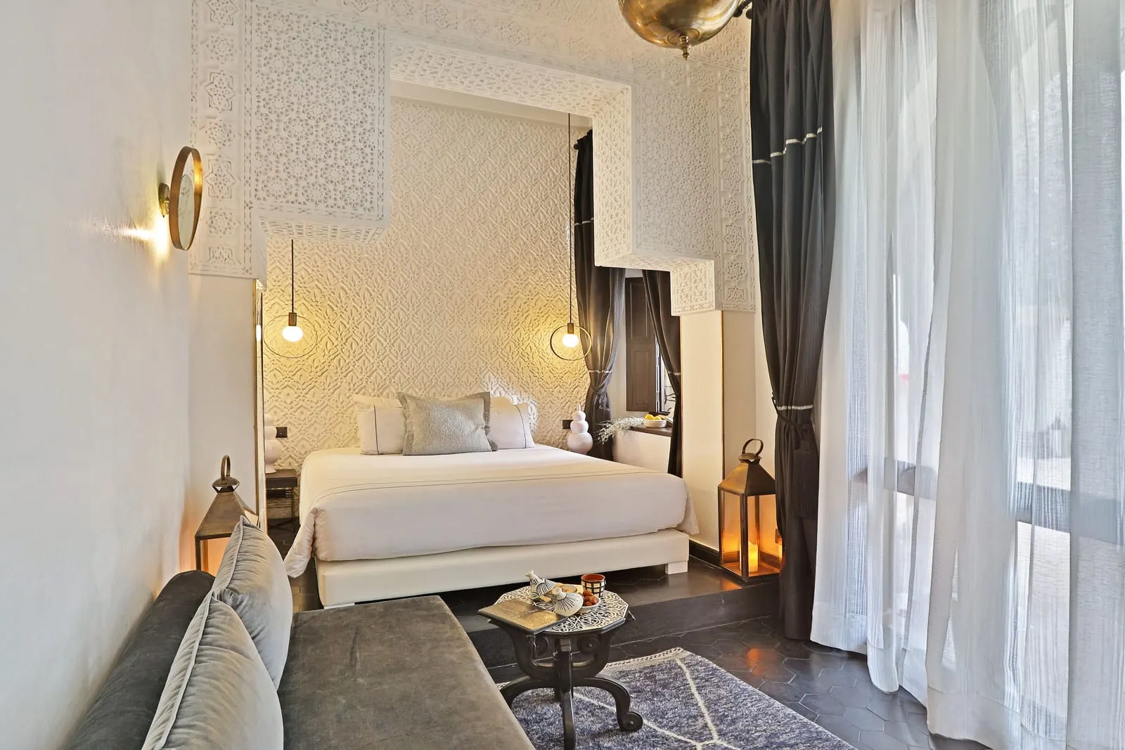 Suite Touch DéCo at Dar Kandi: a white bed on a raised platform under carved white plasterwork, with a grey sofa and lanterns