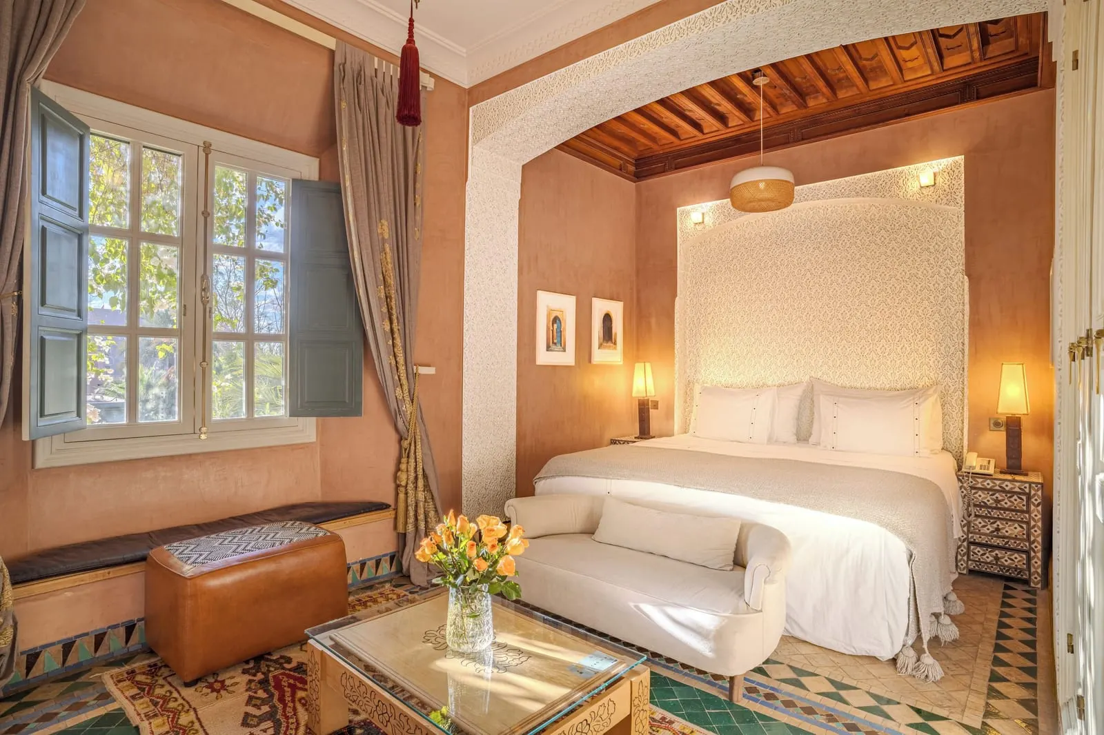 Serail Double at Dar Rhizlane: a large double bed under a carved cedar ceiling, with a cream sofa at its foot and a window seat beside shuttered windows