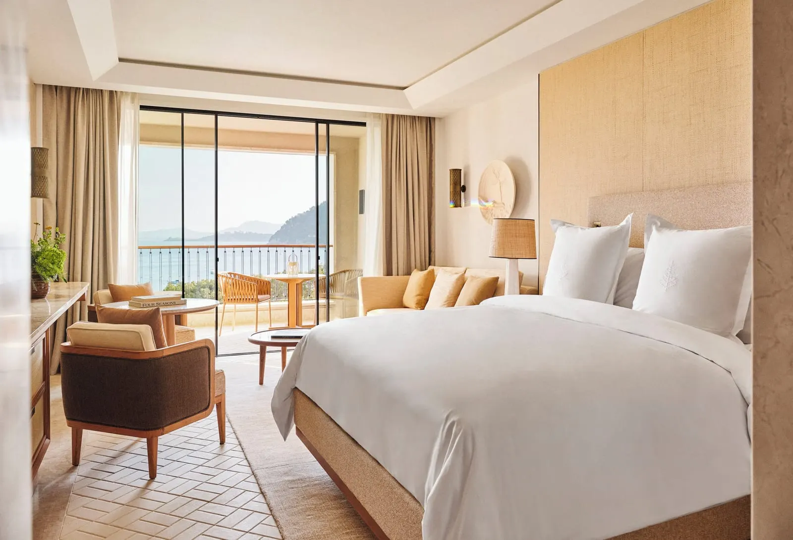Sea-View Room at Four Seasons Resort Mallorca at Formentor: a king bed with a woven headboard, a sofa and an armchair, and glass doors onto a terrace with the sea and hills beyond the balustrade