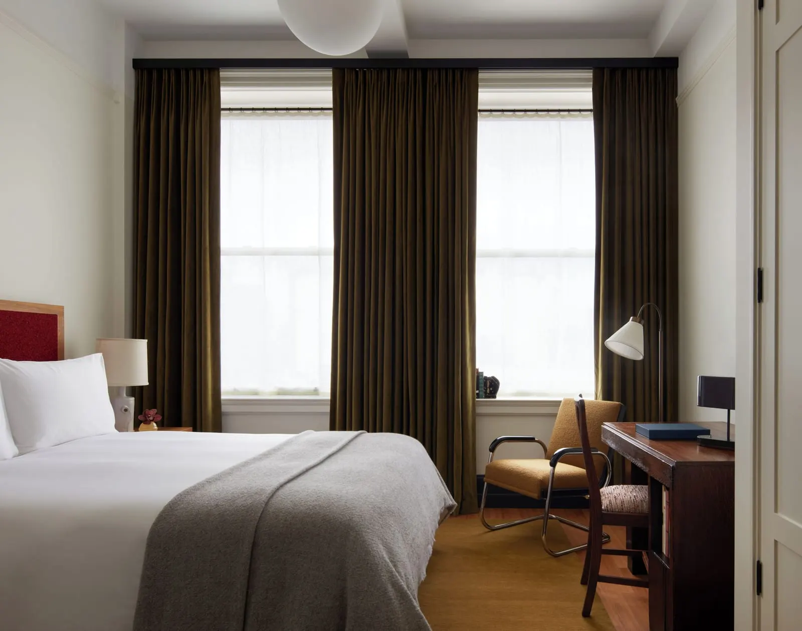 Classic Queen at Nine Orchard: a bed with a grey wool throw in front of two tall windows with long olive curtains, an armchair and a wooden desk beside them
