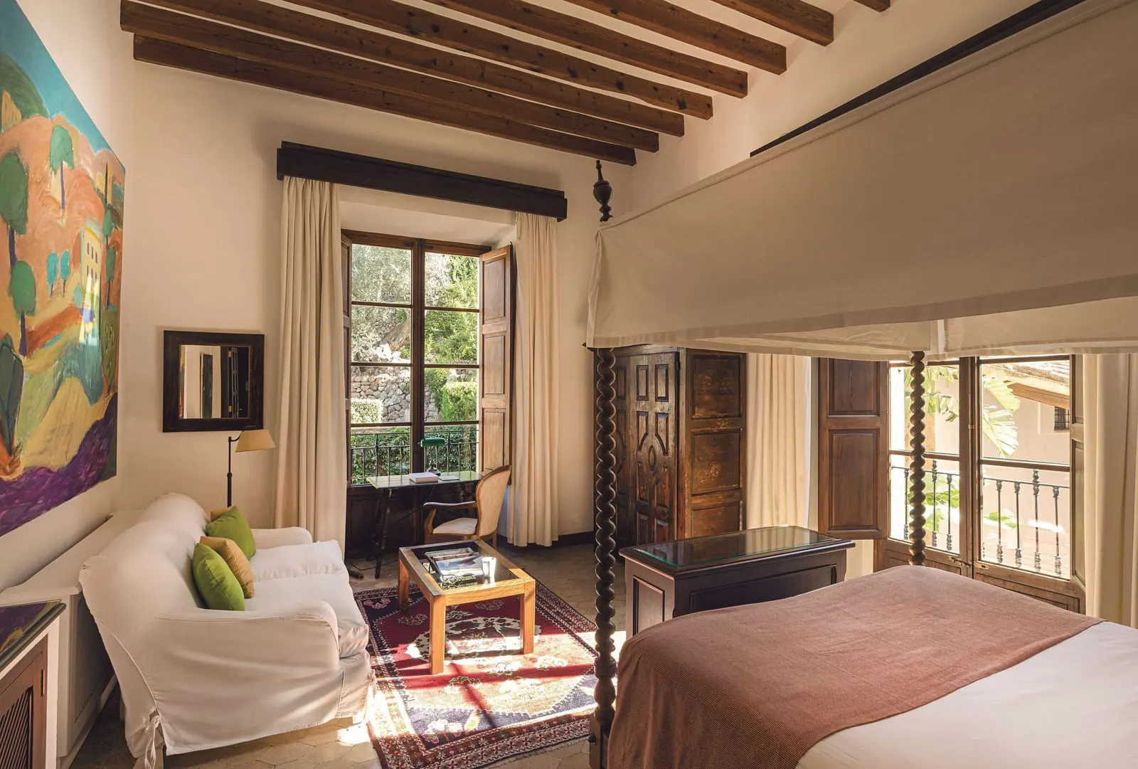 Superior Double Room at Belmond La Residencia: a four-poster bed under wooden beams, a white sofa on a red rug and a desk by a window with an iron balcony rail
