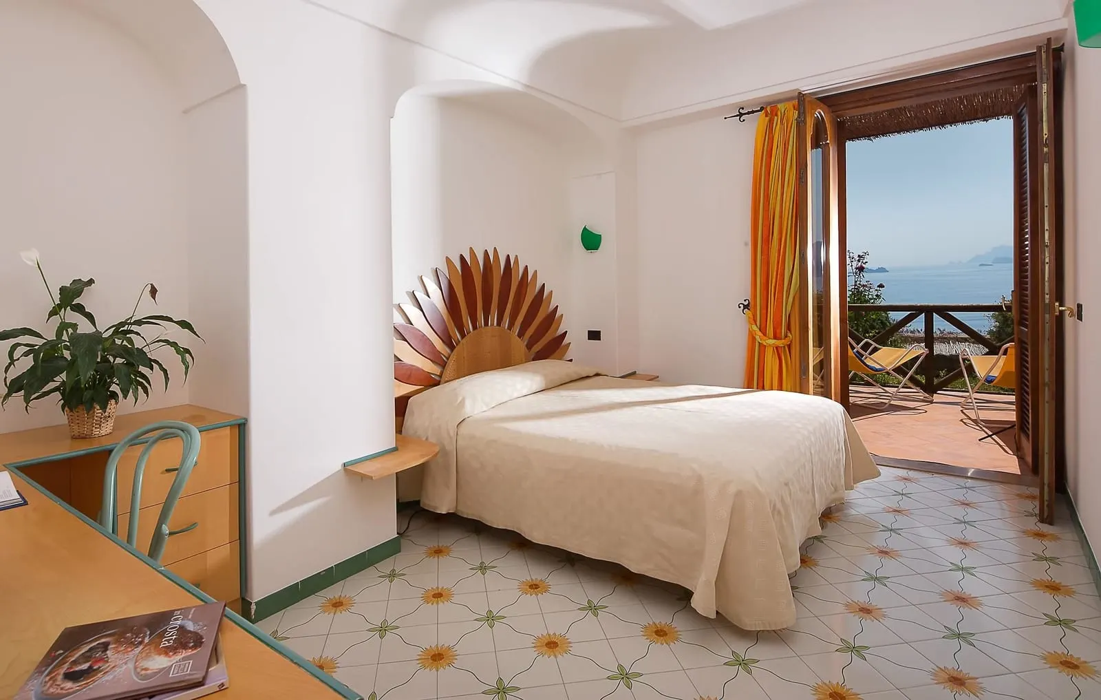 Superior Room with sea view terrace all'Hotel Tramonto d'Oro: un letto matrimoniale con testiera in legno a ventaglio su un pavimento piastrellato dipinto a mano, con porte aperte su una terrazza con sedie pieghevoli e il mare in lontananza.