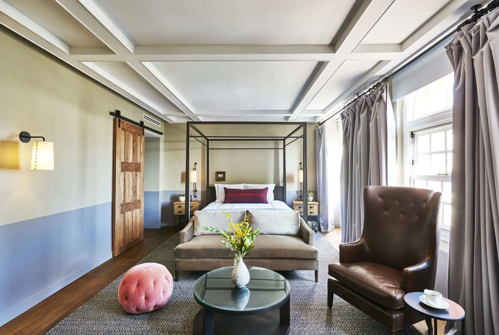 Tower Suite im The Hollywood Roosevelt: ein Himmelbett mit einem Sofa am Fußende, ein Ledersessel, ein rosa Pouf und eine Holztür im Scheunentor-Stil, unter einer Kassettendecke
