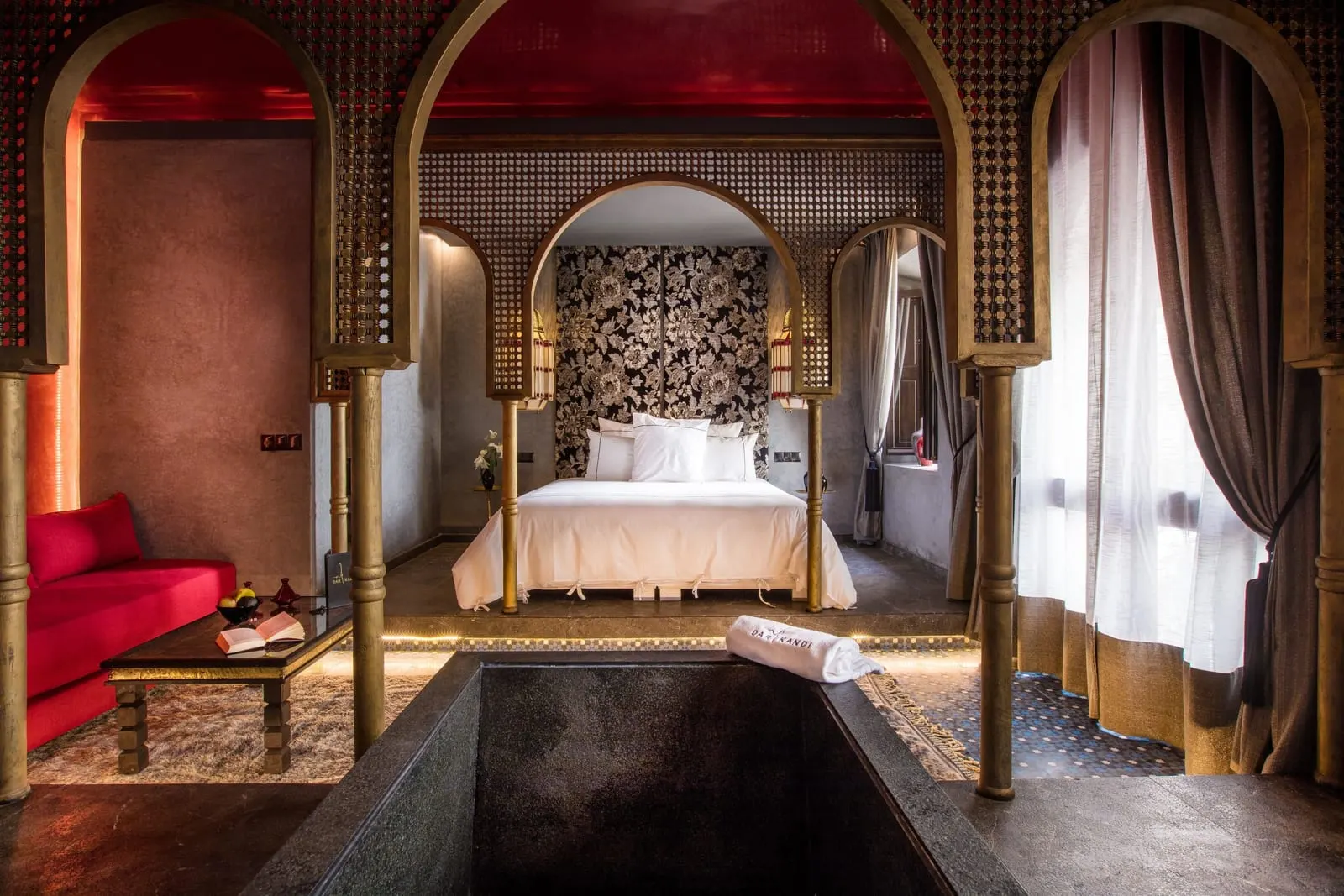 Suite Arch at Dar Kandi: a white bed framed by carved wooden arches, with a red sofa and a sunken bath in the foreground