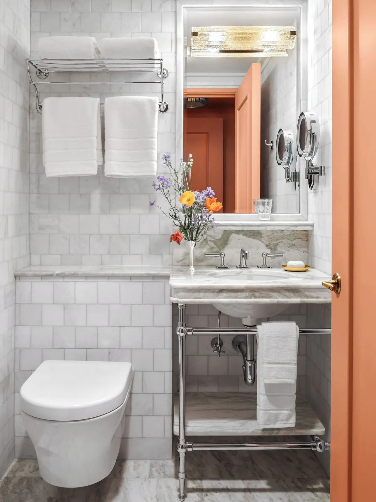 The NoMad King at The Fifth Avenue Hotel: a marble bathroom with a pedestal basin on a chrome stand, a towel rail above a wall hung toilet, and a coral painted door