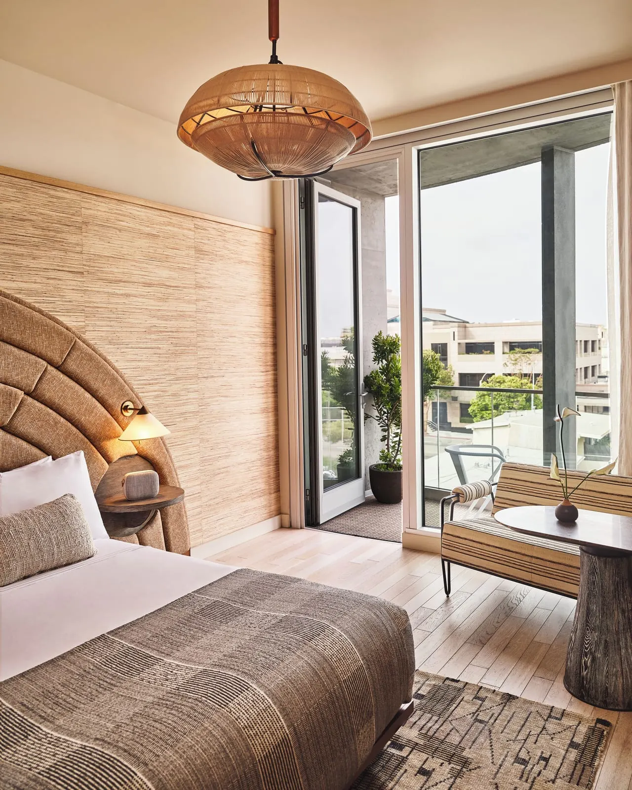 Premier Balcony King im Santa Monica Proper Hotel: ein Bett neben einer Pendelleuchte und einem gestreiften Stuhl, mit offenen Fenstertüren zu einem Balkon mit einem Baum im Topf