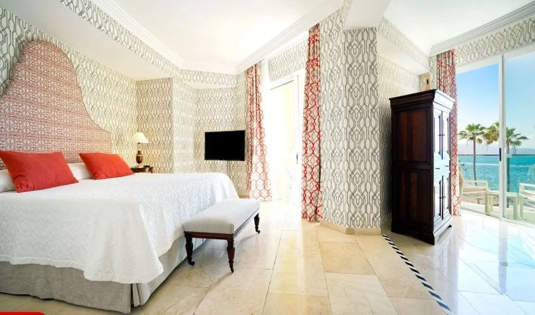 Sea View Suite at Bull Reina Isabel & Spa: a double bed with a tall patterned headboard and red cushions, a bench at its foot, a dark wooden wardrobe and a glass door to a balcony with palms and the sea beyond