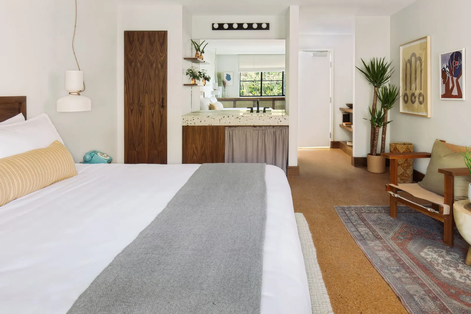 The Deluxe King at Silver Lake Pool & Inn: the foot of a bed with a grey runner, a vanity alcove with a terrazzo counter, a tall walnut door and an armchair on a patterned rug