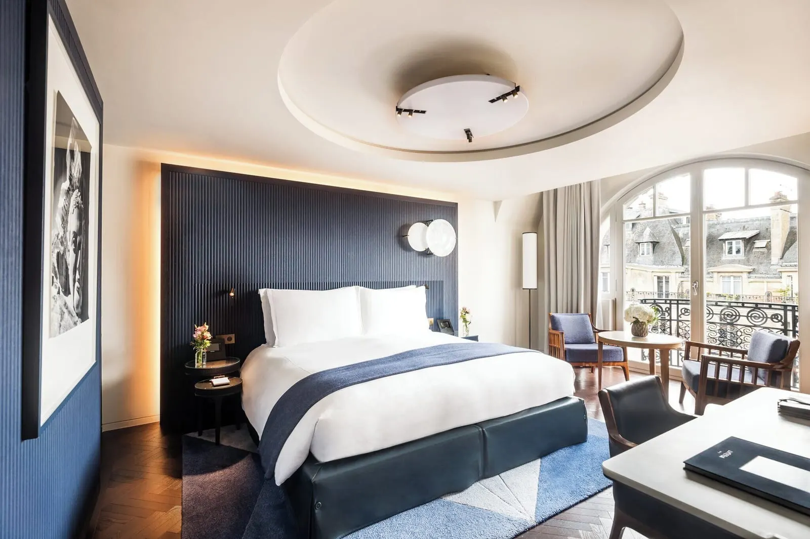 Deluxe Balcony Room at Mandarin Oriental Lutetia, Paris: a double bed against a dark blue fluted headboard, a writing desk, and two armchairs and a round table by arched French windows onto a wrought-iron balcony