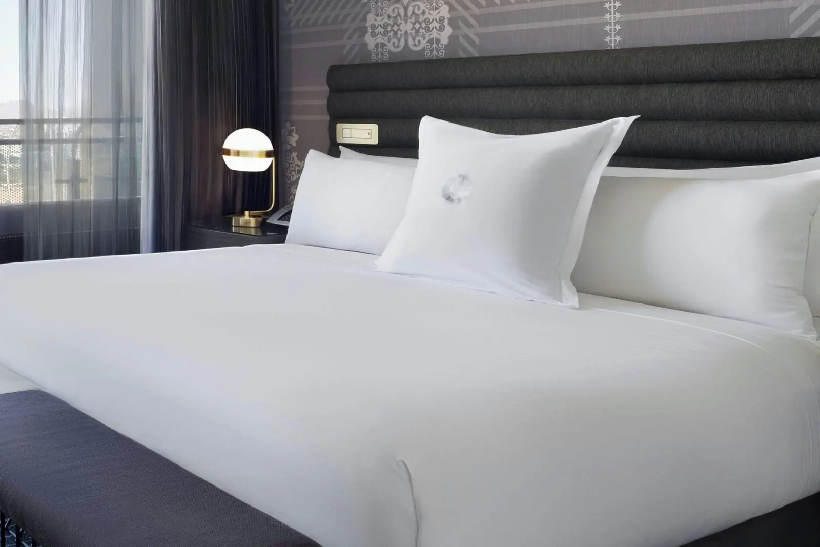 Terrace Studio King Fountain View at The Cosmopolitan of Las Vegas: a king bed with a padded charcoal headboard, a brass globe lamp on the bedside table and a long upholstered bench at its foot