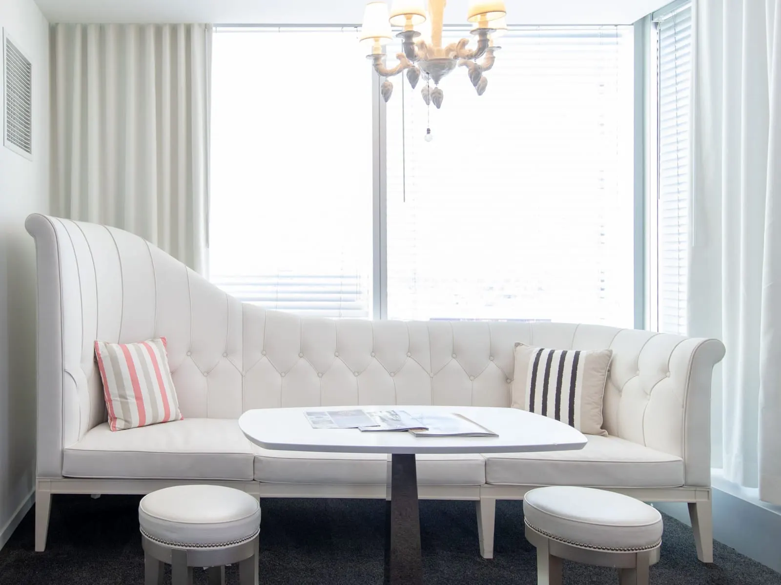 Alexandria King Suite at SAHARA Las Vegas: a white tufted banquette with a table and two stools in a corner with windows on two sides, under a chandelier