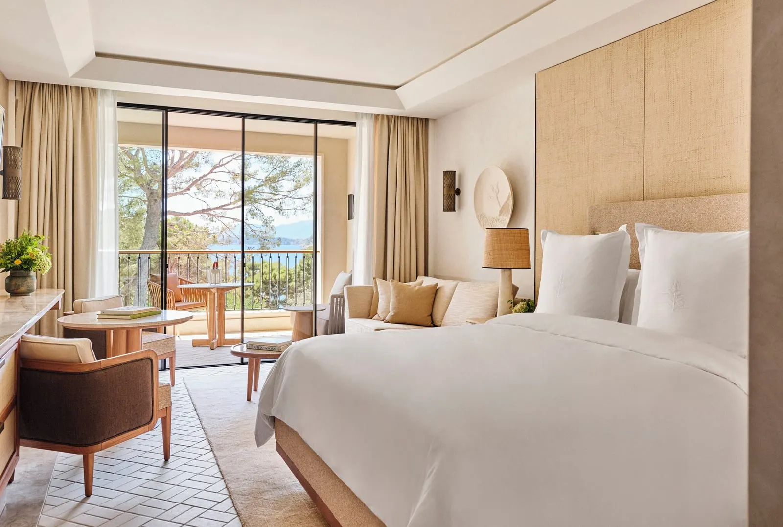 Pine-View Room im Four Seasons Resort Mallorca at Formentor: ein Kingsize-Bett mit einem Kopfteil aus Flechtpaneelen, ein Sofa und ein runder Tisch und Glastüren zu einer Terrasse zwischen Kiefern