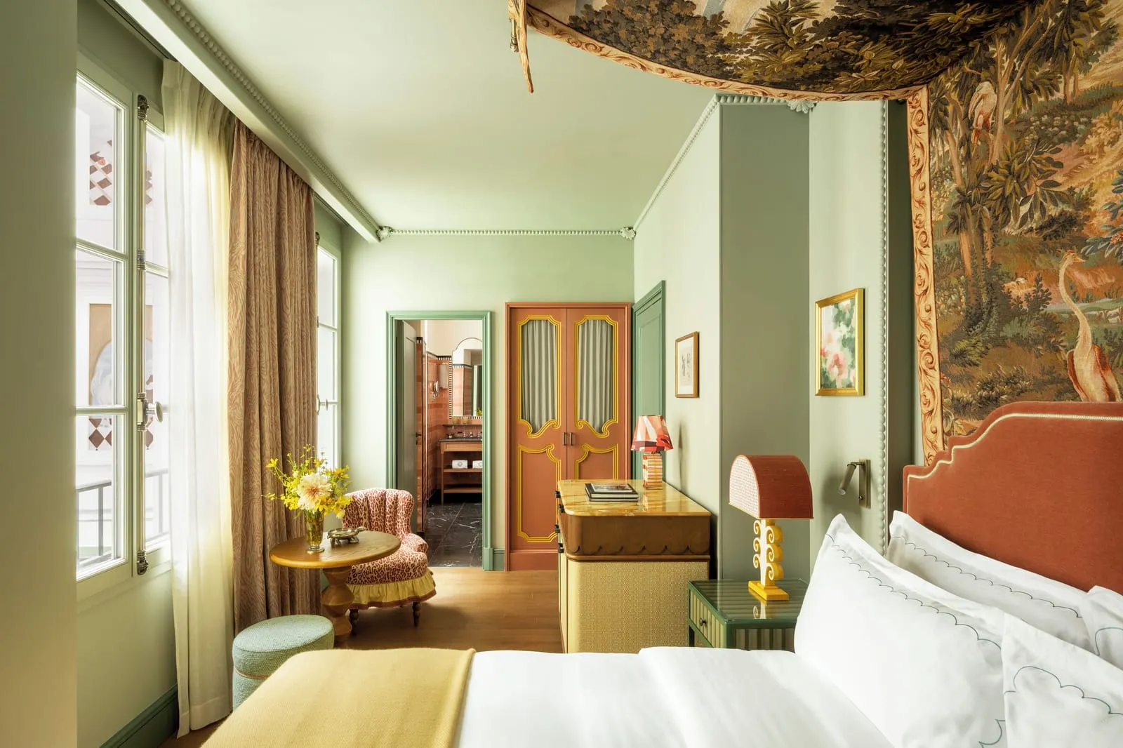 Classic Room at Le Grand Mazarin: a bed with a rust velvet headboard under a tapestry canopy, a leopard-print armchair by tall windows and the tiled bathroom through an open door