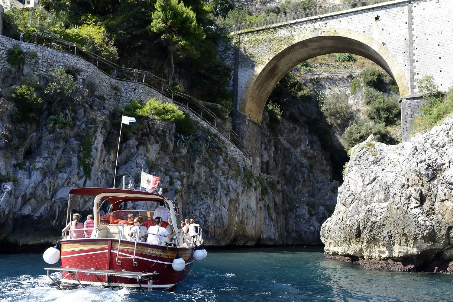 A red wooden motor boat with guests aboard entering the Furore fjord, under the arched bridge of the coast road
