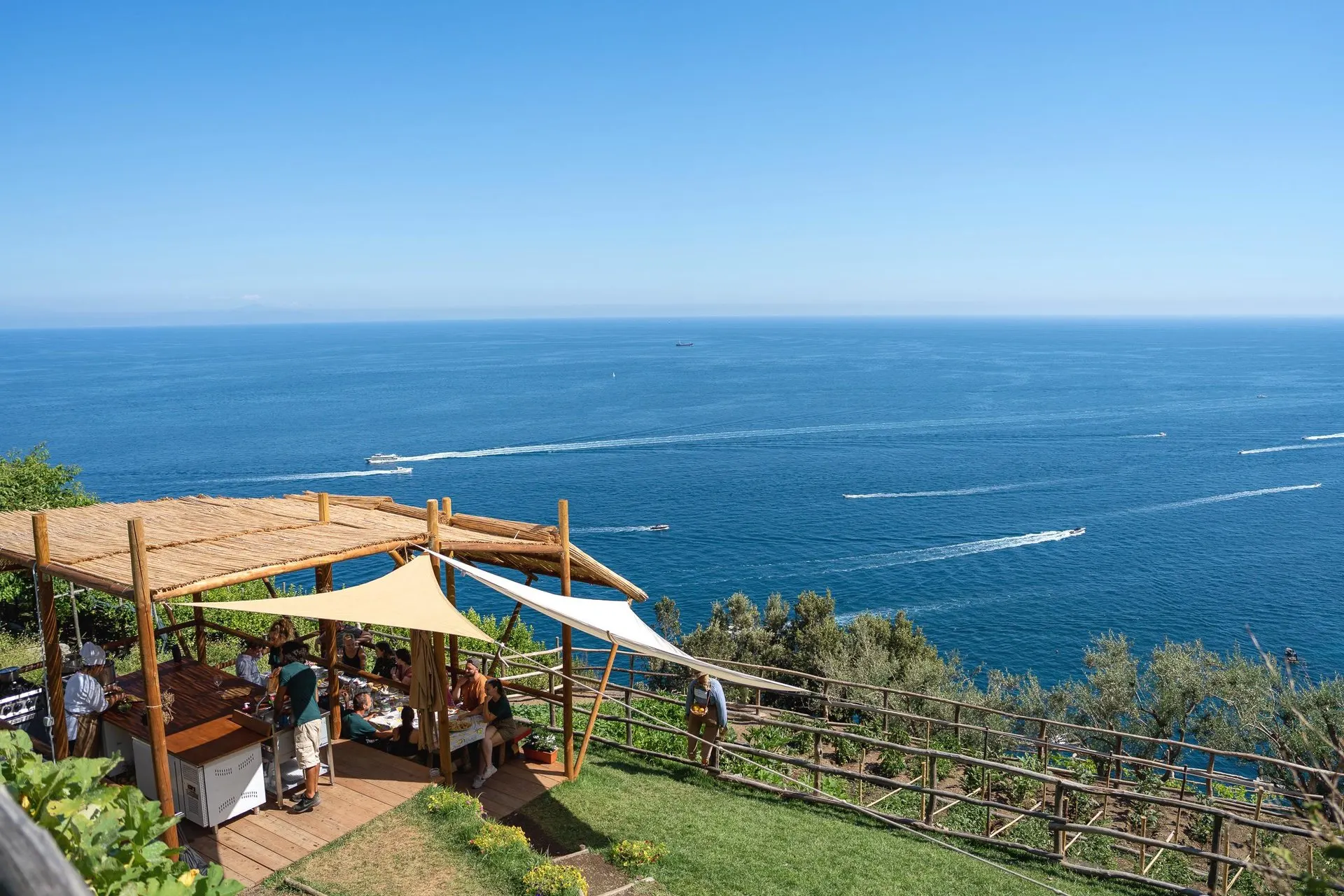 A class eating under a reed-roofed pergola on a terraced garden high above the sea, with boats crossing the bay below
