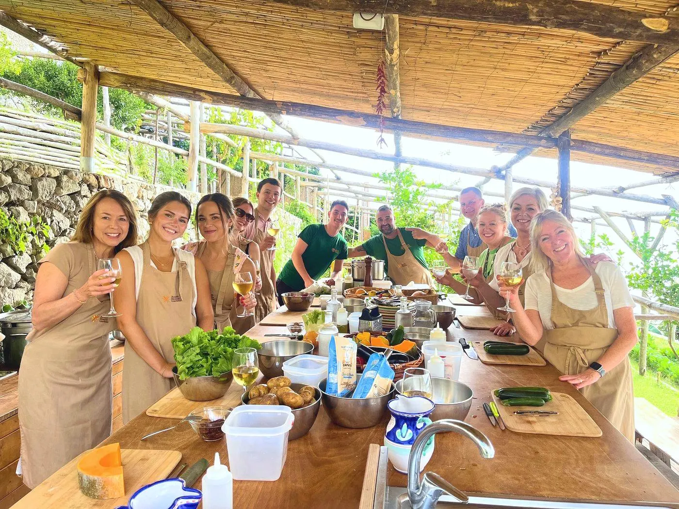 A cooking class in aprons raising glasses of white wine at a long worktable of vegetables under a reed pergola