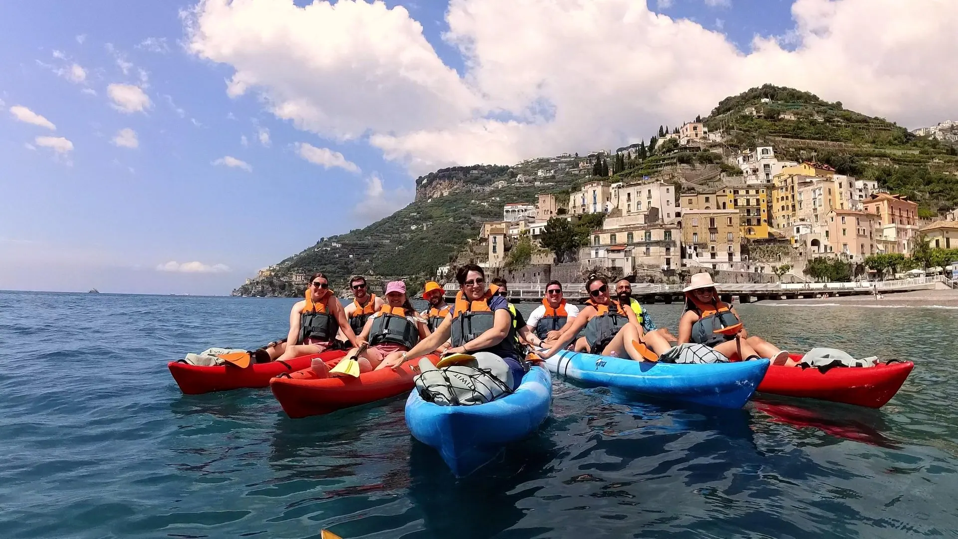 A group in red and blue sit-on-top kayaks off Minori, with the town's seafront and terraced hillside behind