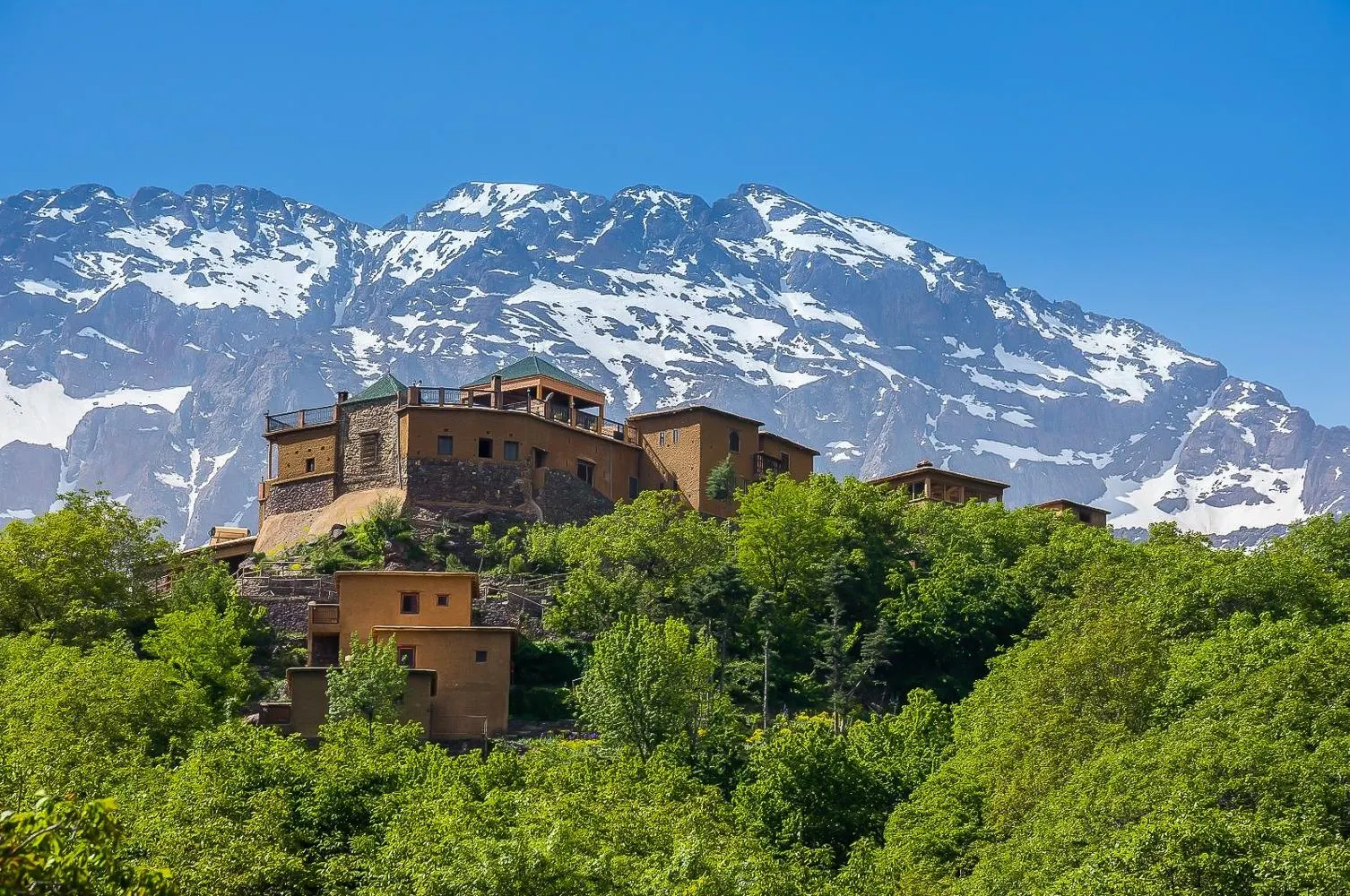 Earth-built houses on a wooded hilltop above Imlil, with the snow-streaked ridges of the Toubkal massif behind