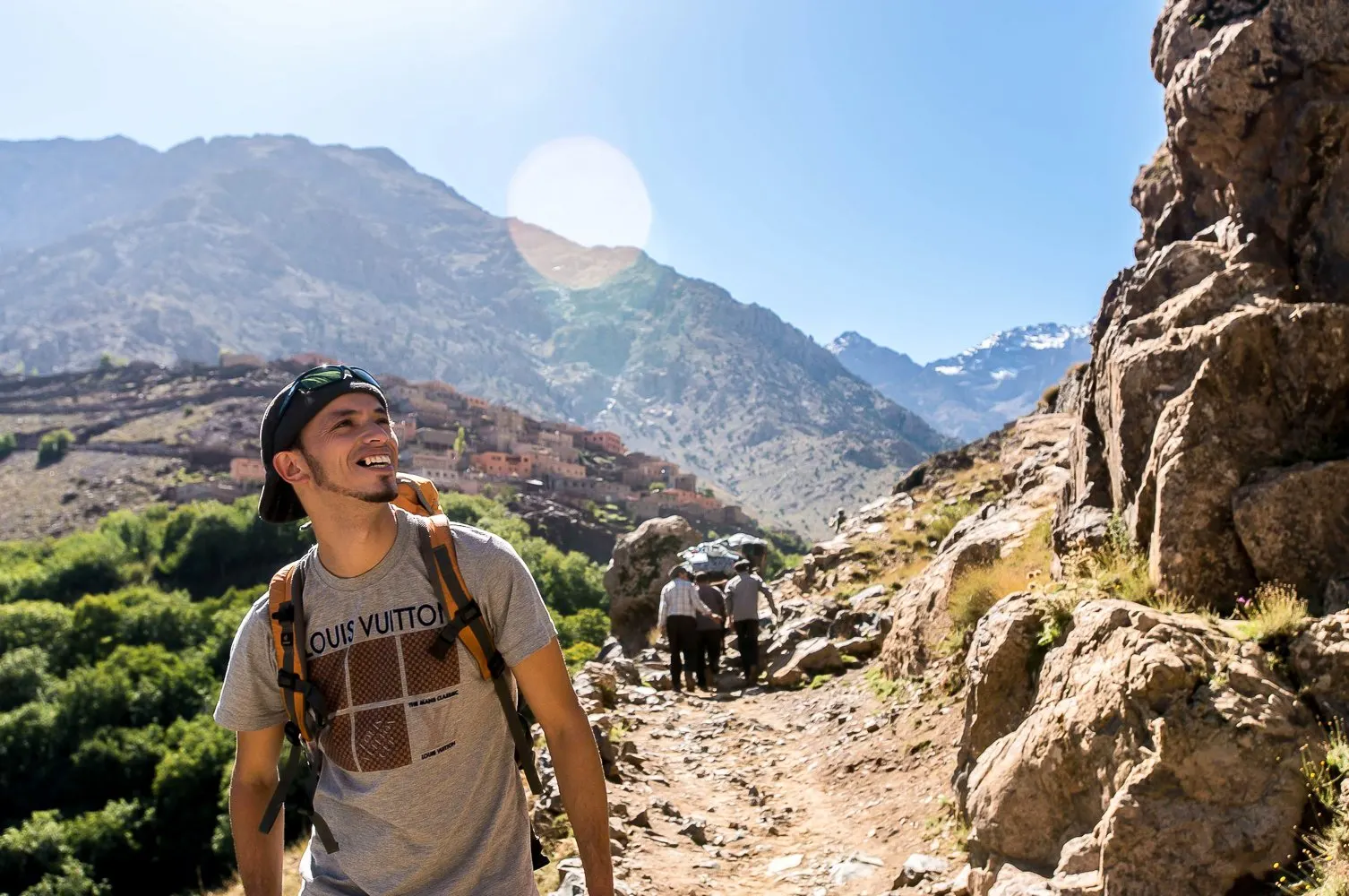 A walker with a day pack on a stony path in the High Atlas, with a terraced village and a snowy peak behind
