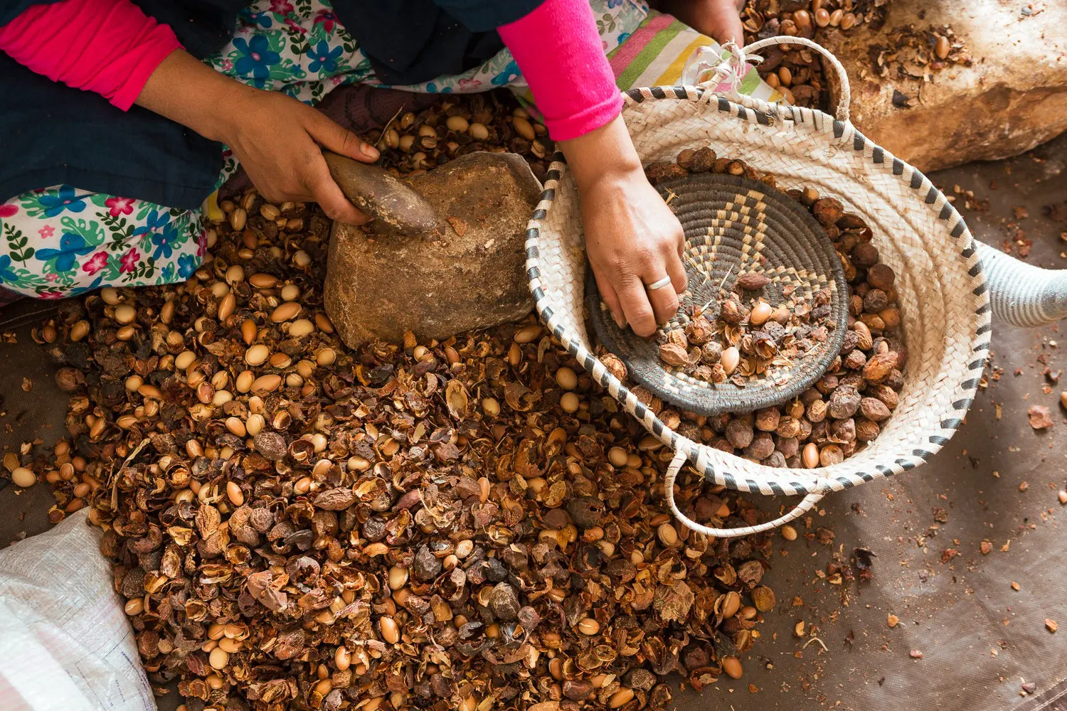 A woman cracking argan nuts with a stone beside a basket of kernels