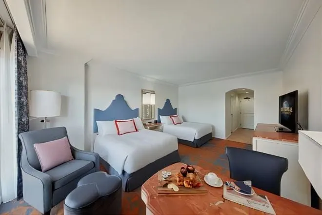 Club 2 Queen Room at Loews Portofino Bay Hotel: two queen beds with blue upholstered headboards, an armchair and ottoman by the window and a marble-topped table, with a television on the dresser