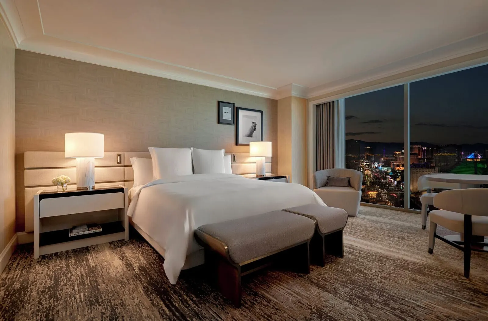 Strip View Room at Four Seasons Hotel Las Vegas: a king bed with a two-part bench at its foot, lit bedside lamps, and an armchair and a round table by the window at night