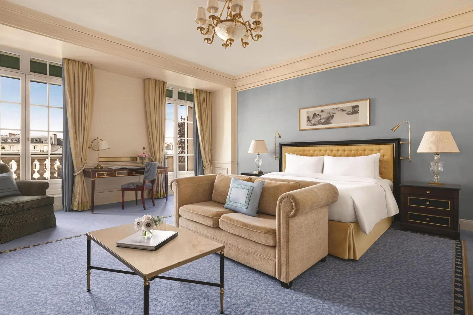 Terrace Room at Shangri-La Paris: a king bed with a gold headboard against a blue wall, a velvet sofa and a stone-topped coffee table, with a writing desk between tall French windows