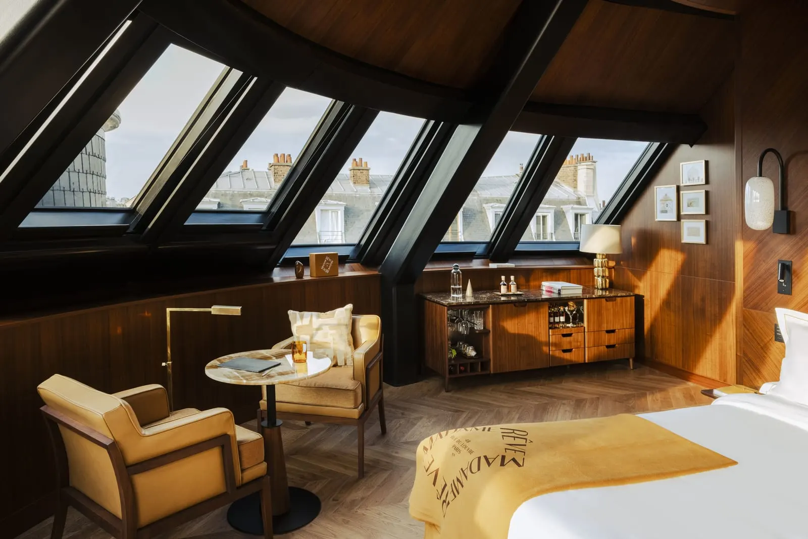 Suite Pigeonnier at Hôtel Madame Rêve: a curved run of sloping atelier windows under a curved timber ceiling, onto grey mansard roofs and chimney pots, with two armchairs, a round stone table, a minibar cabinet and the foot of the bed