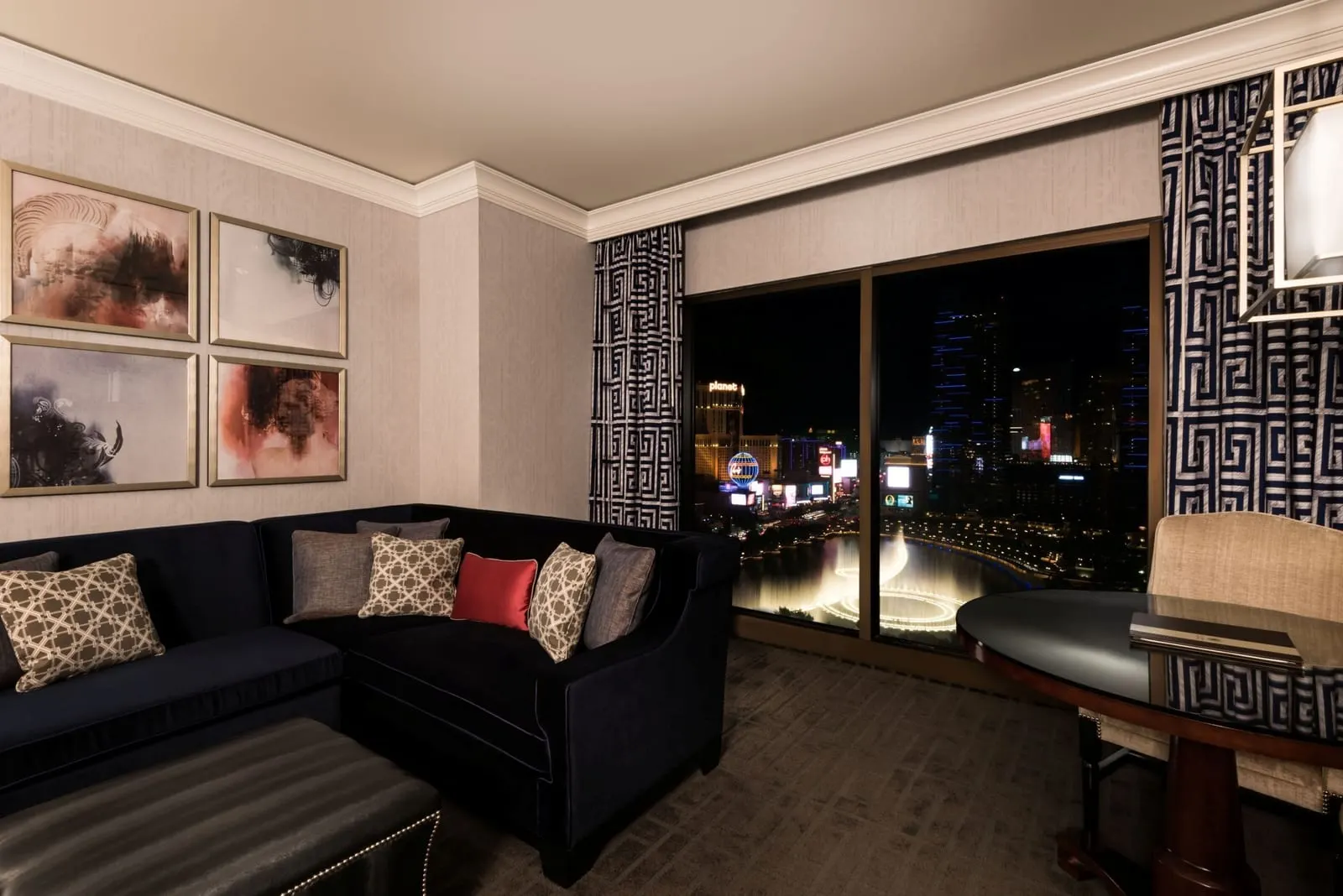 Augustus Fountain View Room at Caesars Palace, as it was before the 2026 remodel: a sectional sofa and a round table beside a window onto the Bellagio fountains at night