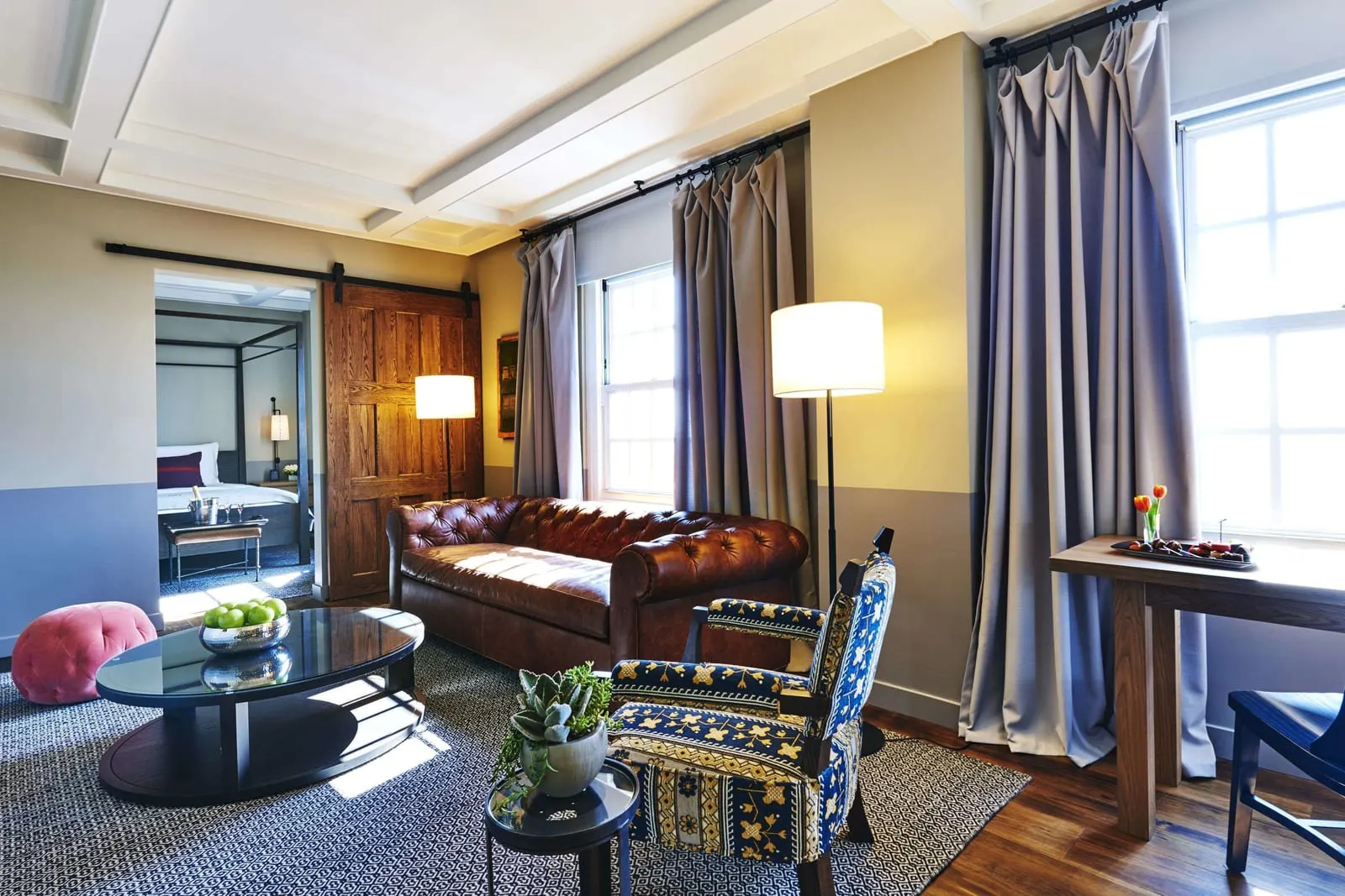 Blossom Suite im The Hollywood Roosevelt: ein Wohnzimmer mit einem hellbraunen Chesterfield-Ledersofa, einem gemusterten Sessel und einem ovalen Couchtisch, mit einer offenen Tür im Scheunentor-Stil zu einem Himmelbett