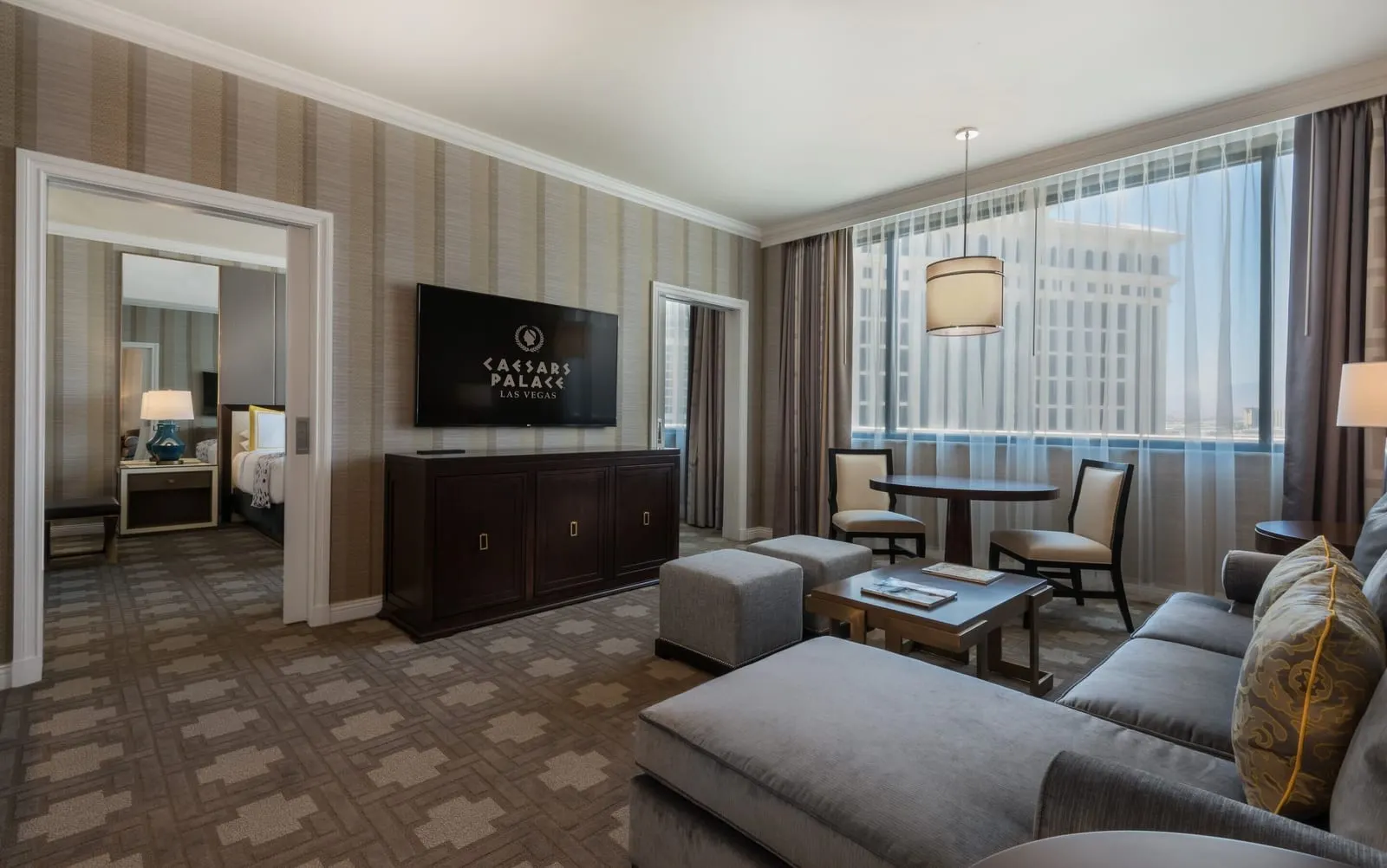 Julius Signature Level Suite im Caesars Palace: ein Wohnzimmer mit einem grauen Ecksofa, einem Tisch für zwei am Fenster, einem Fernsehschrank und einer Tür zum Schlafzimmer