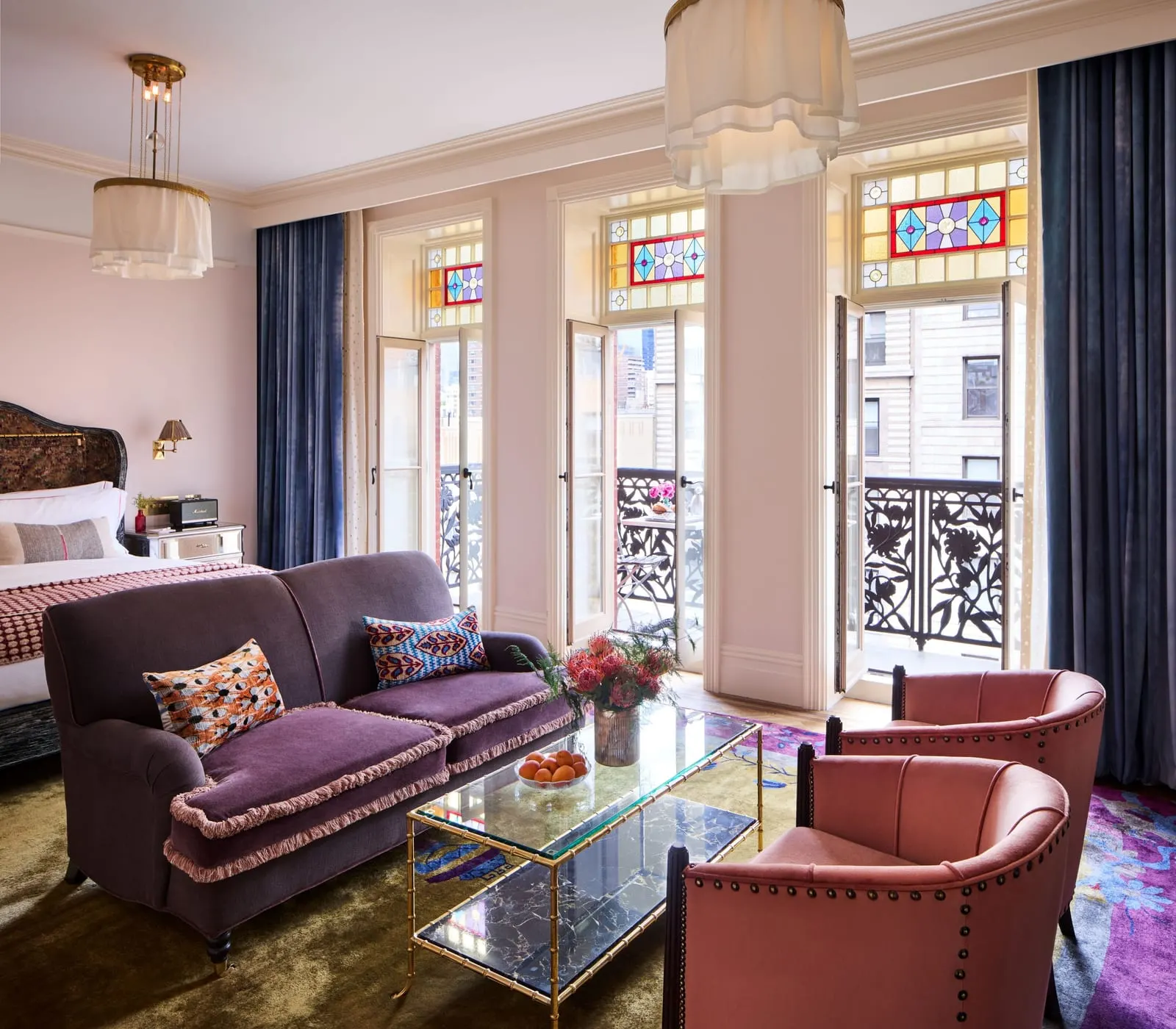Premier Chelsea at Hotel Chelsea: a purple sofa, two pink armchairs and a glass coffee table in front of three tall doors with stained-glass panels above them, with the bed at the left edge