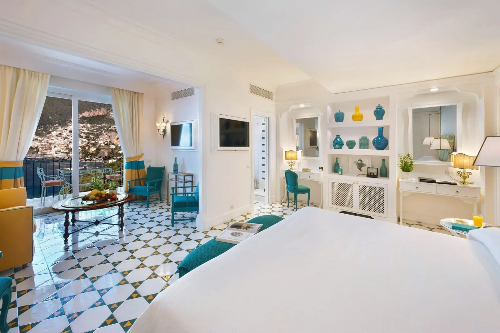 Signature Room at Il San Pietro di Positano: a large bed facing a sitting area and shelves of turquoise ceramics, with doors to a terrace