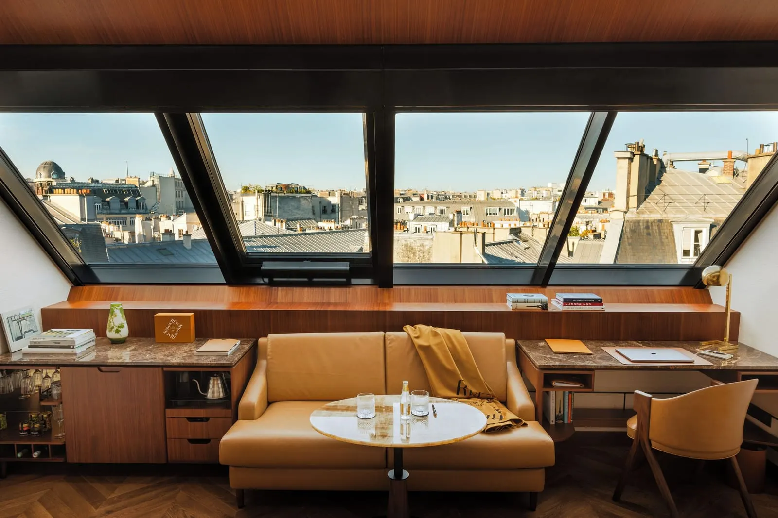Deluxe Atelier, Paris View, at Hôtel Madame Rêve: a leather sofa, a round marble table and a writing desk beneath a wall of sloping atelier windows onto grey Paris rooftops and chimneys