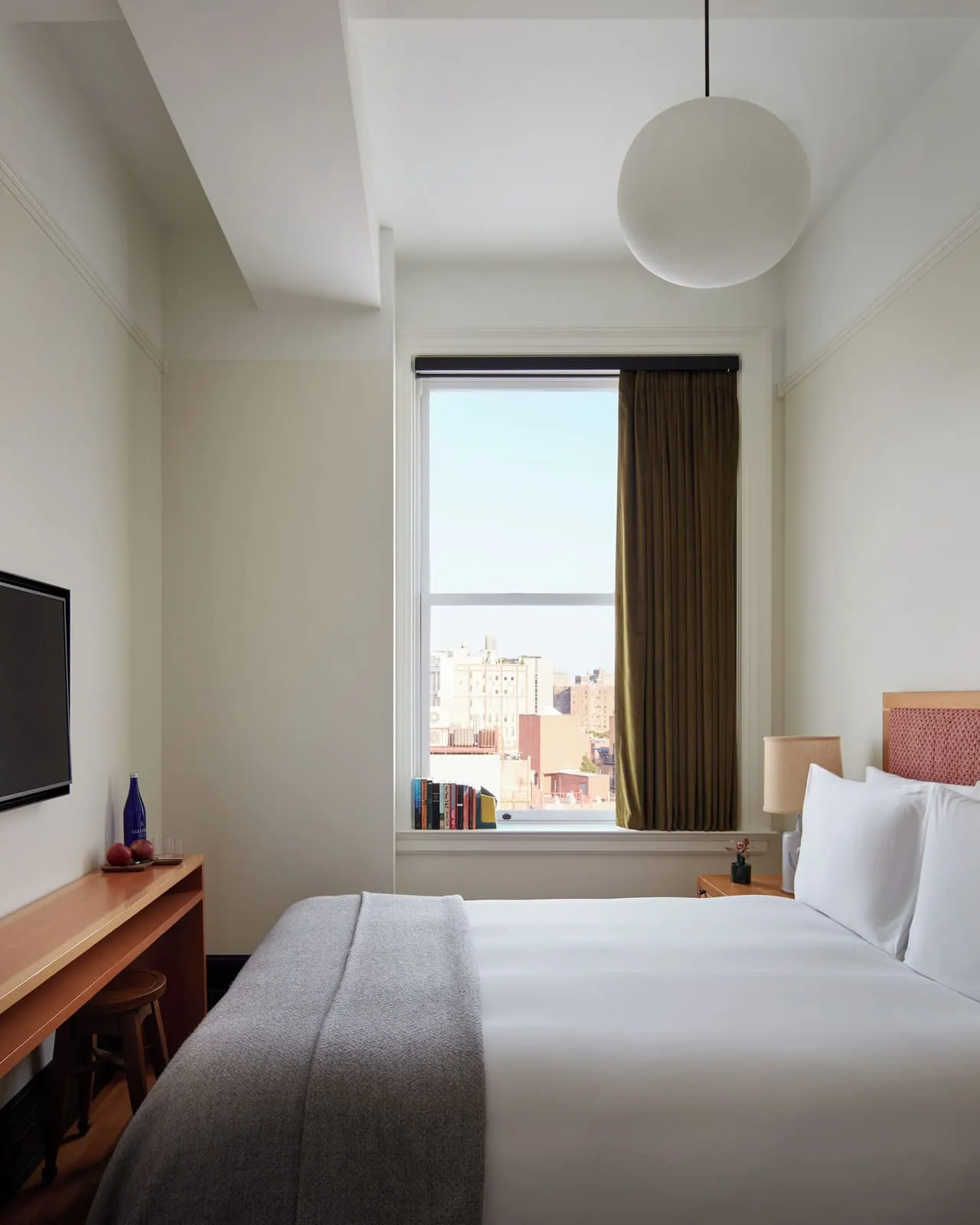 Nook at Nine Orchard: a bed with white linen and a grey wool throw in a narrow room, with a tall window at the far end, a pendant globe light and a slim wooden shelf desk with a wall-mounted television
