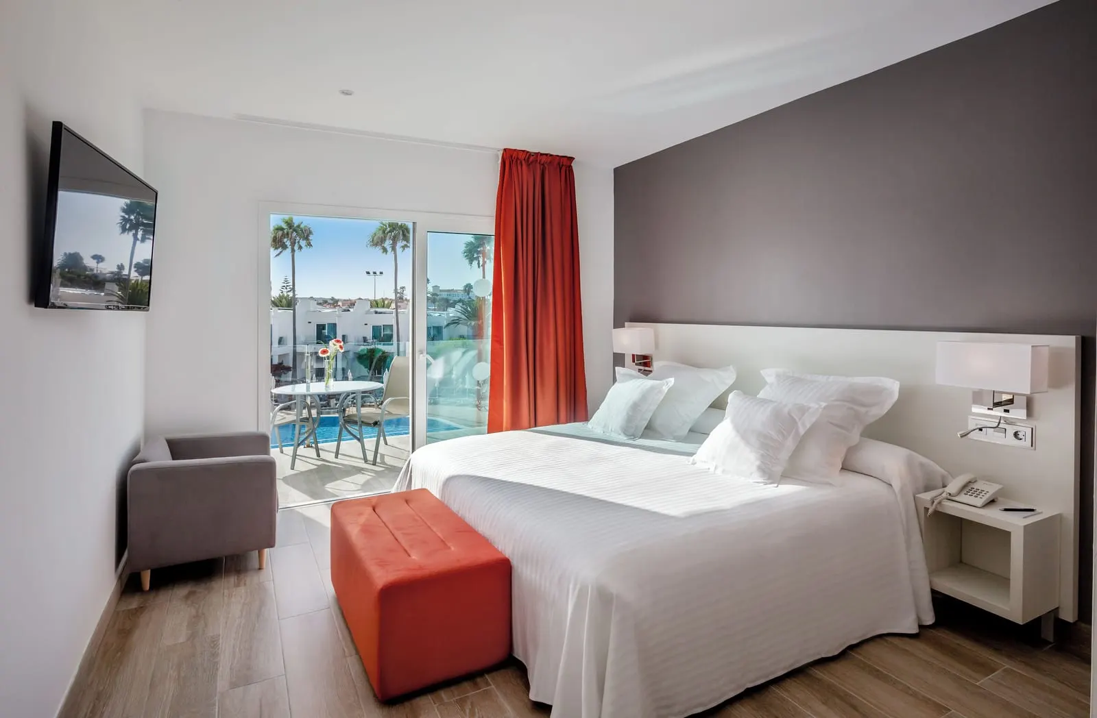 Double at Barceló Corralejo Sands: a bed with white linen, an orange footstool, an armchair and glass doors onto a balcony.