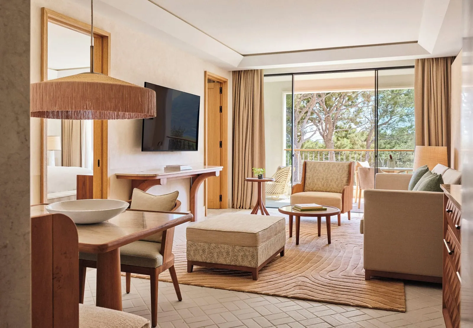 Grand Pine-View Suite im Four Seasons Resort Mallorca at Formentor: ein Wohnzimmer mit einem Esstisch im Vordergrund, einem Sofa, einem Sessel und einem Polsterhocker auf einem gemusterten Teppich, einem Wandfernseher und Glastüren zu einer Terrasse zwischen Kiefern