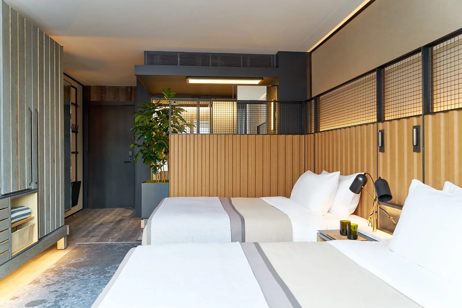 Dumbo 2 Beds im 1 Hotel Brooklyn Bridge: zwei Doppelbetten mit grauen und hellbraunen Überwürfen vor einer Kopfteilwand aus geripptem Holz, ein hoher Kleiderschrank aus Holz links und eine große Pflanze am Eingang