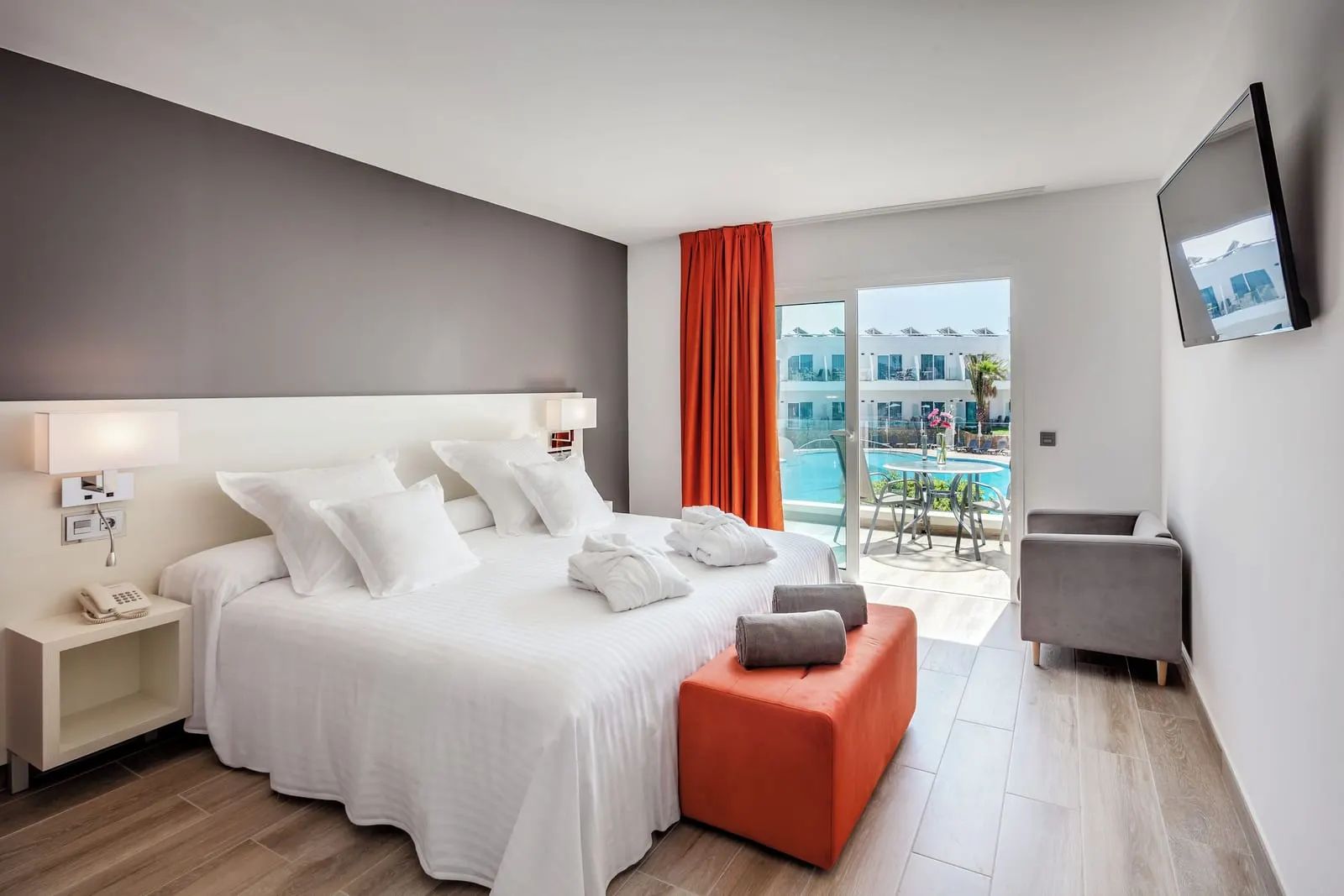Deluxe at Barceló Corralejo Sands: a large bed with an orange footstool, an armchair and glass doors onto a balcony.