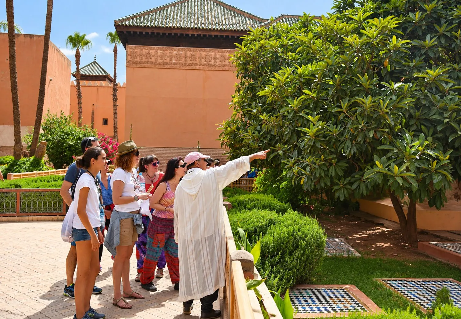A guide in a white djellaba points out the garden of the Saadian Tombs to a small group, with the tiled roof of the mausoleum behind
