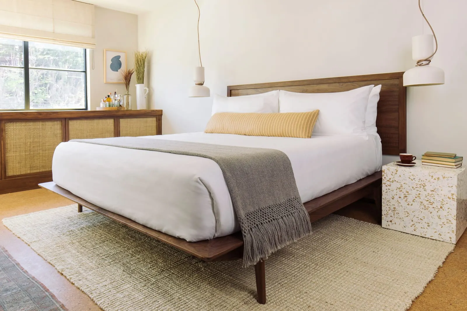 The Deluxe King View at Silver Lake Pool & Inn: a king bed with a grey throw and a yellow striped bolster cushion, a terrazzo bedside table and a window on the left