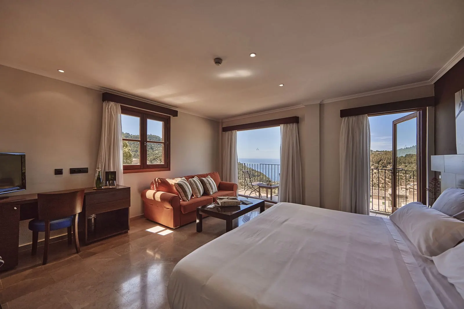 Sea Junior Suite at Maristel Hotel & Spa: a large bed, an orange sofa with a coffee table and a desk, and balcony doors onto hills with the sea in the distance