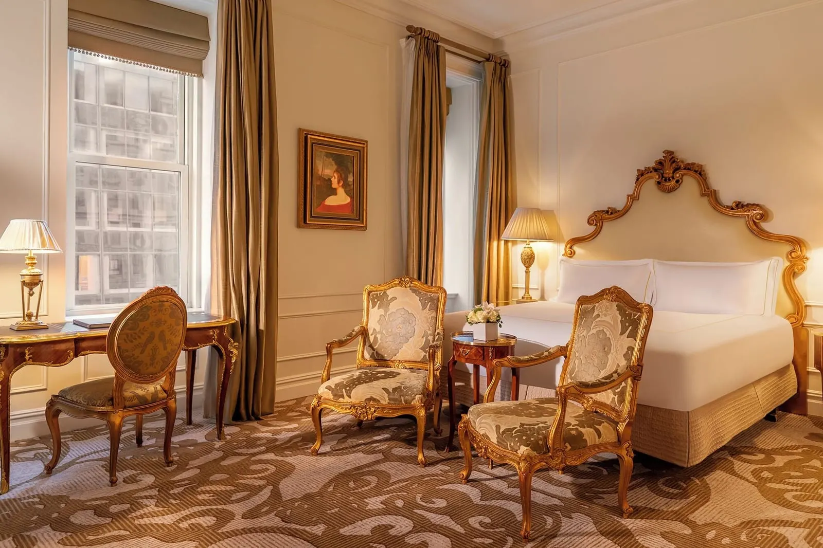 Deluxe King at The Plaza: a king bed with a gilded headboard, two gilt armchairs and a writing desk beside a tall window, in warm light